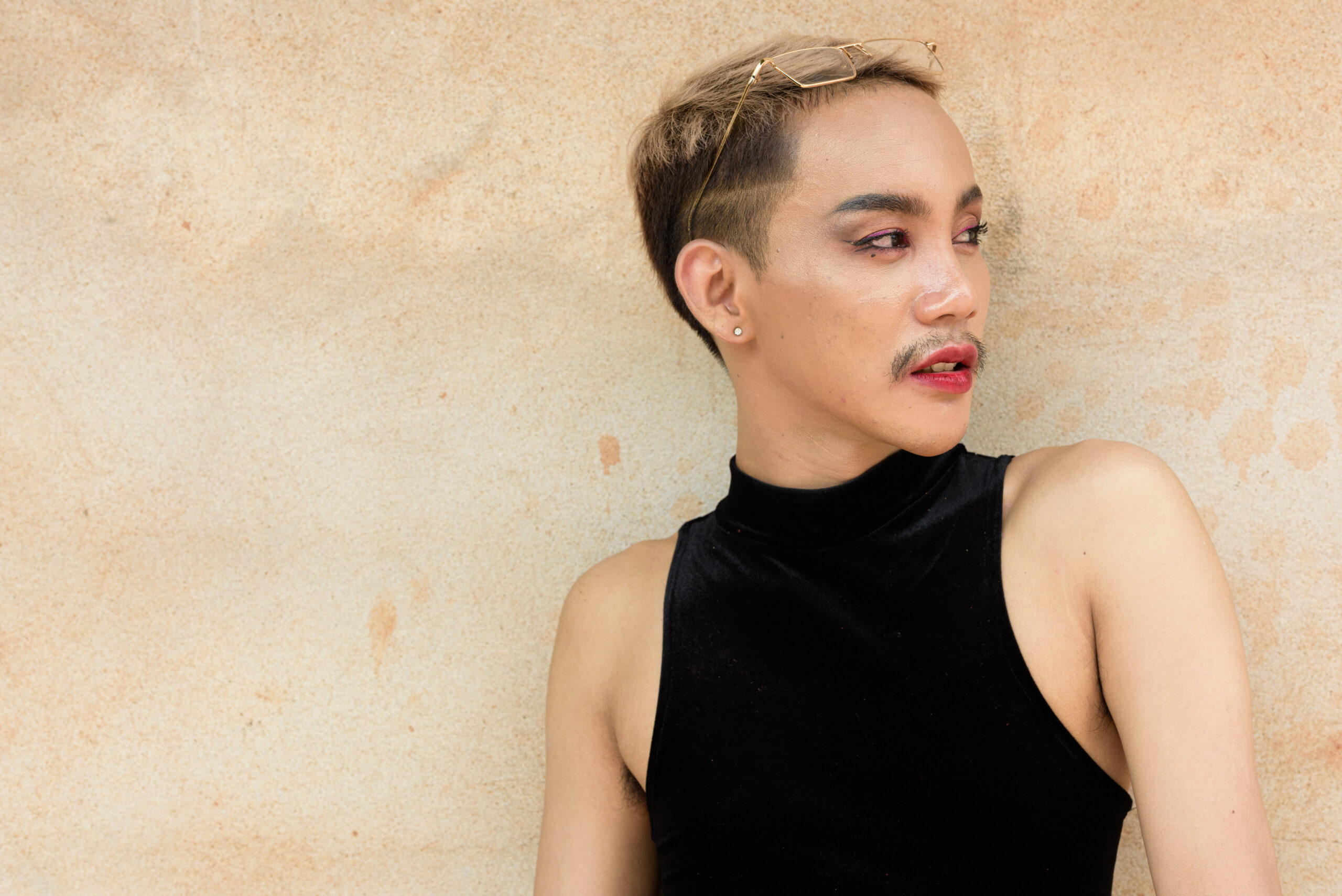 Portrait of beautiful Asian queer LGBT community supporter man with mustache wearing lipstick and jumpsuit against beige wall background